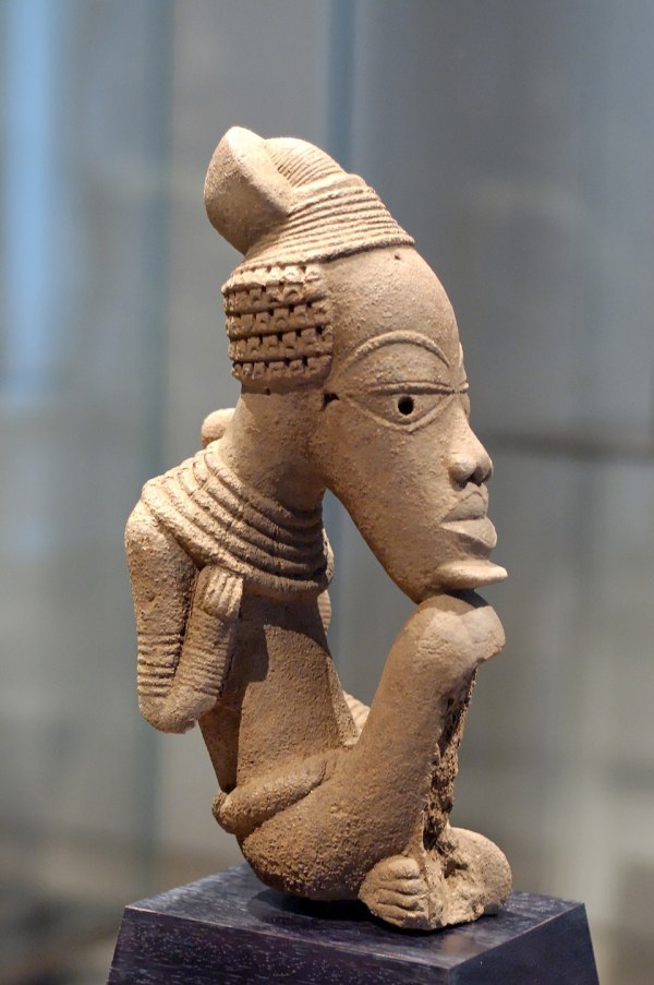 Photograph of a seated Nok terracotta sculpture depicting a figure resting its chin on its knee; an ancient Nigerian terracotta artwork from between about 500 BCE and 500 CE now in the Louvre Museum.