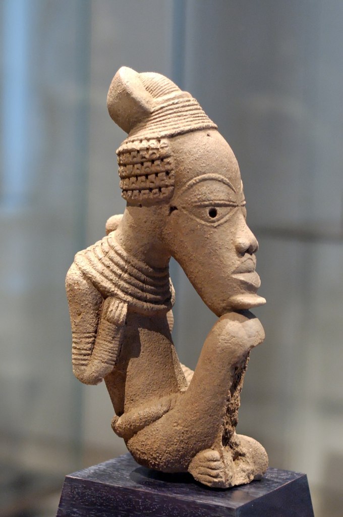 Photograph of a seated Nok terracotta sculpture depicting a figure resting its chin on its knee; an ancient Nigerian terracotta artwork from between about 500 BCE and 500 CE now in the Louvre Museum.