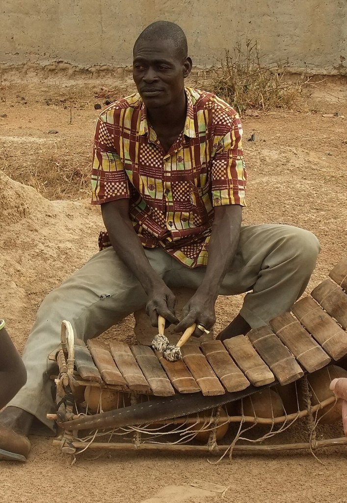 West African griot playing a balafon, a traditional wooden xylophone that accompanies oral storytelling and musical heritage.