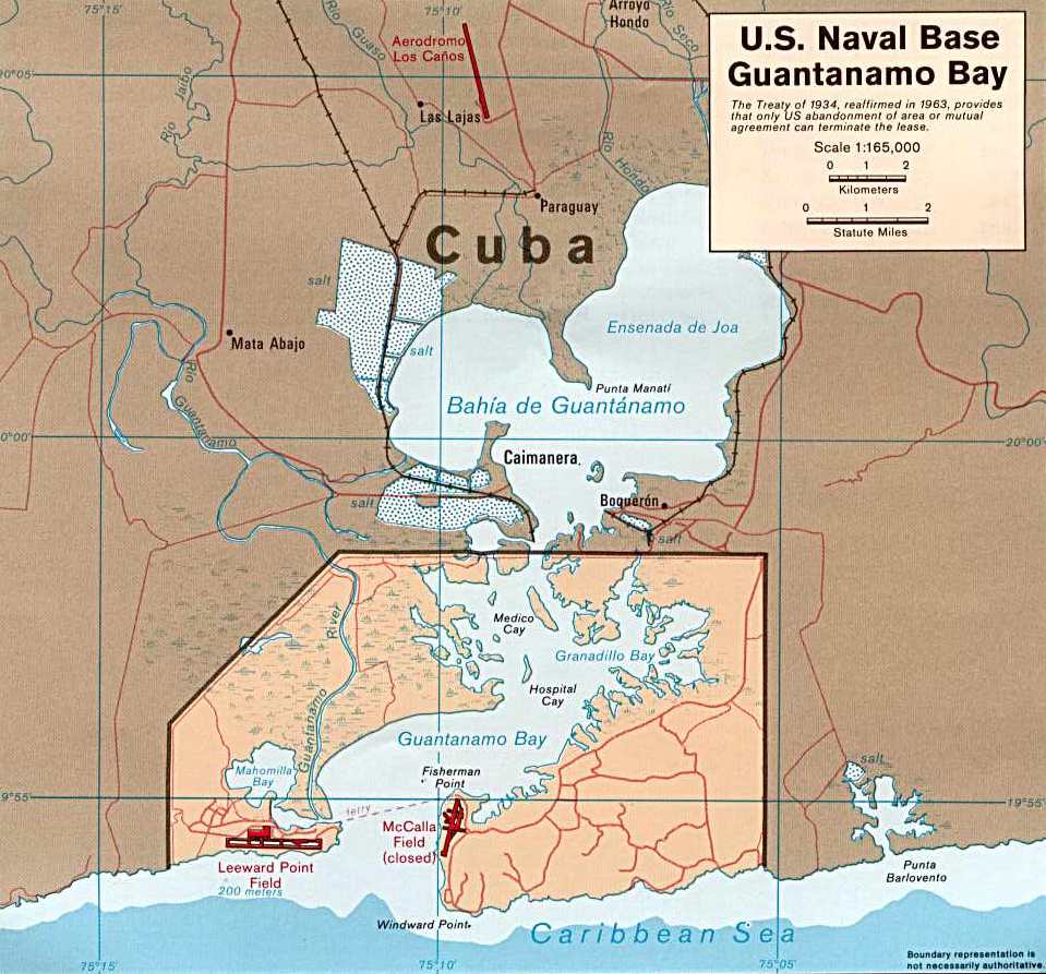 Aerial view of the United States Naval Station at Guantánamo Bay, Cuba, showing the base’s shoreline, runways, buildings, and dock facilities at the southeastern edge of the bay.