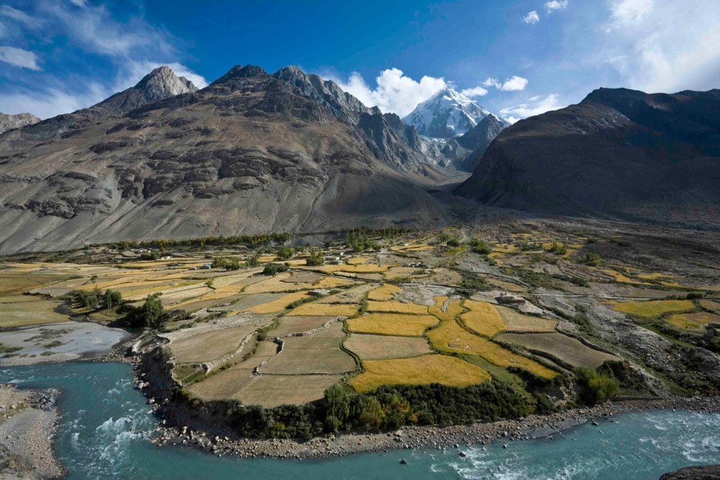 A remote village in the Wakhan Corridor of Afghanistan, with small fields along a river valley and rugged mountains rising in the background under a partly cloudy sky.