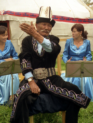 A Kyrgyz manaschi (oral storyteller) performing the epic Manas in Karakol, Kyrgyzstan, wearing traditional clothing and gesturing expressively.